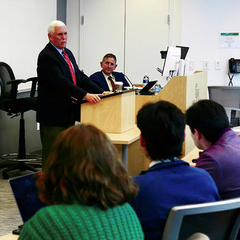 A man in a suit stands behind a lectern while another man in a suit watches.